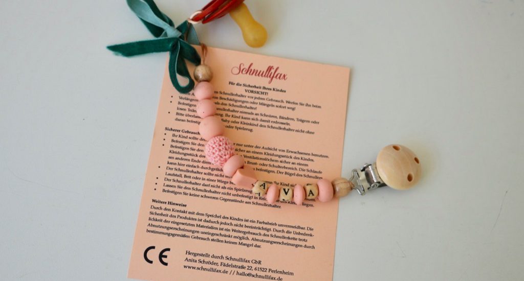 Warnhinweise Schnullerketten: Beipackzettel mit allen wichtigen Infos im Postkarten-Format; die Schnullerkette ist daran mit einer Schleife befestigt.