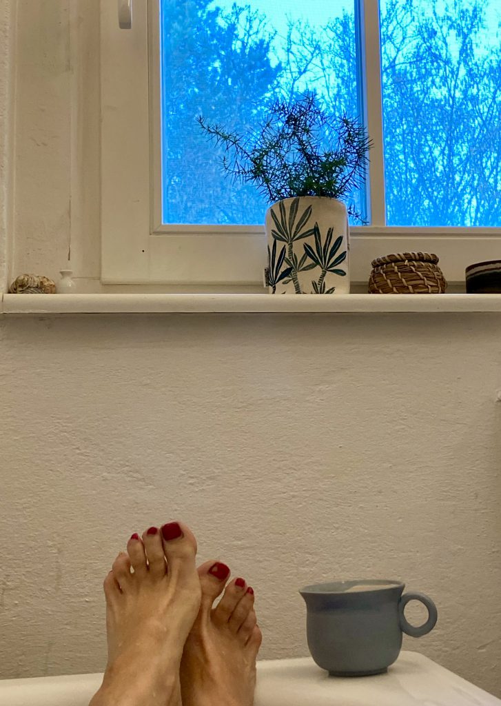 Füße auf Badewannenrand vor kleinem Fenster, mit Teetasse auf dem Badewannenrand