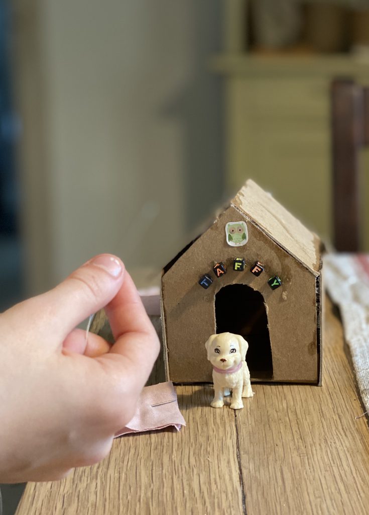 kleine Hundehütte aus Pappe auf Holztisch mit weißem Hündchen davor, Kinderhand mit Nähfaden und Stoff davor