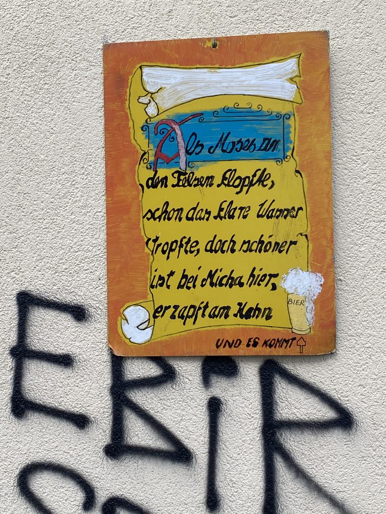 Ein perfekter Tag in Berlin ist für manche an Bier gekoppelt: Spruch-Tafel an Hauswand: "Als Moses an den Felsen klopfte, schon das klare wasser tropfte. Doch schöner ist's bei Micha hier, er zapft am Krug und es kommt Bier." Darunter vier Graffiti-Buchstaben: EBIR