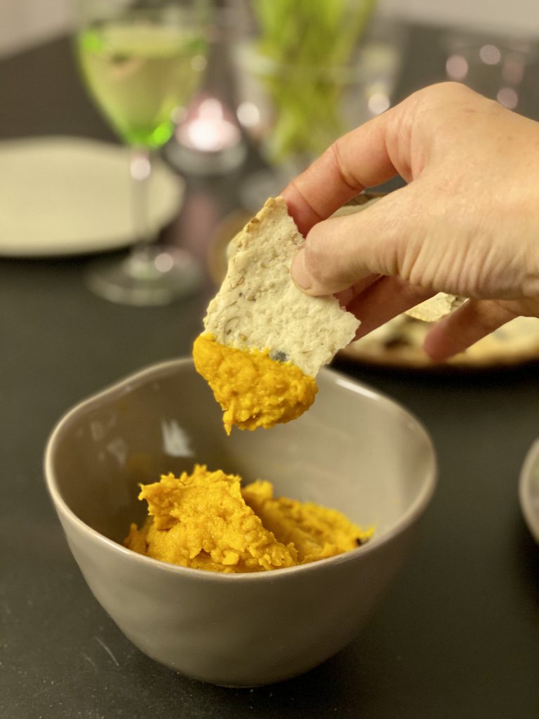 Paleo-Cracker in Hand, mit Dip drauf, vor Weinglas, Schale mit Dip und Teller mit Crackern