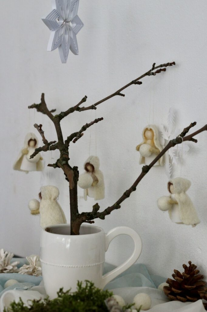 Jahreszeitentisch Puppen (5 Stück) an kahlem Zweig in weißer Tasse, vor weißer Wand
