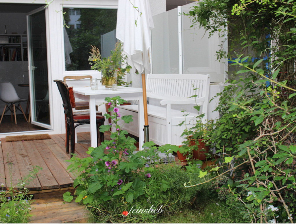 Terrasse aus Holzdielen mit Gartenmöbeln drauf, davor blühen und ranken Blumen