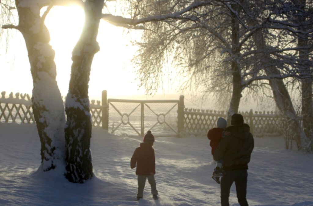 Familie (Vater mit Baby im Arm und zweites Kind) vor Gartentor im Schnee, unter schneebedeckten Bäumen: Ein Spaziergang an Heiligabend/Weihnachten ermöglicht Weihnachten ohne Stress