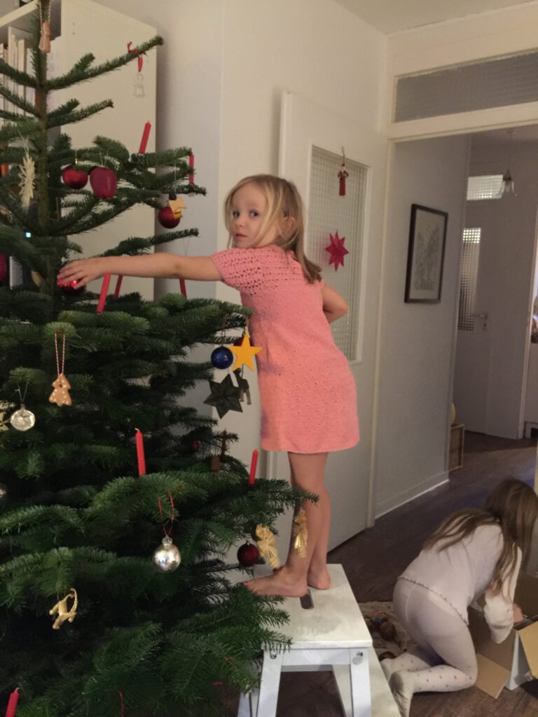 Kind im rosa Kleid schmückt den Weihnachtsbaum, der noch nicht sehr geschmückt ist