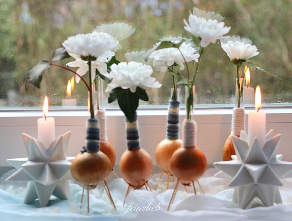 Winterliche Dekoration mit Zwiebel-Vasen auf der Fensterbank, umrahmt mit Kerzen in weißen Leuchtern, alles auf hellblauem Seidentuch