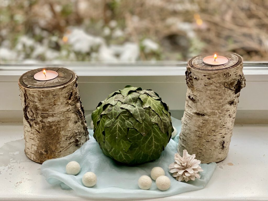 Kugel mit Efeu-Blättern drumrum zwischen Birkenstamm-Teelichthaltern, auf hellblauer Seide, mit weißen Kiefernzapfen und gefilzten Schneekugeln in weiß.