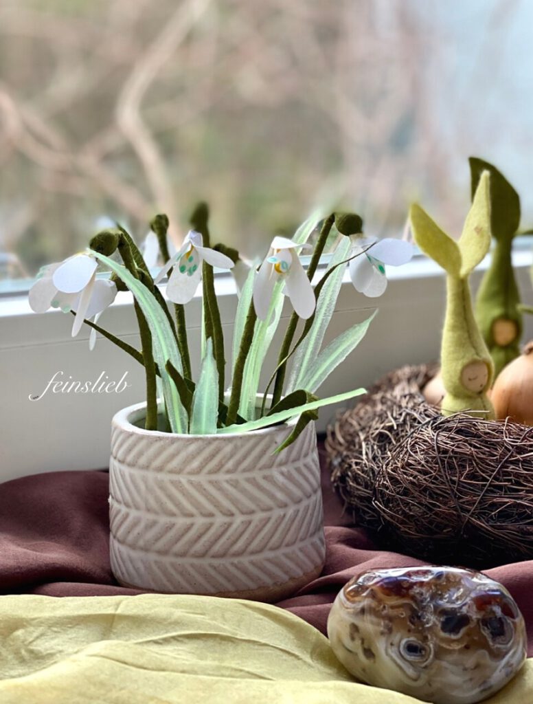Papier-Schneeglöckchen in einer beige-weiß gestreiften Vase auf einer Fensterbank, auf braunem Seidentuch, neben Nest mit Zwiebeln und Sprossen-Blumenkindern