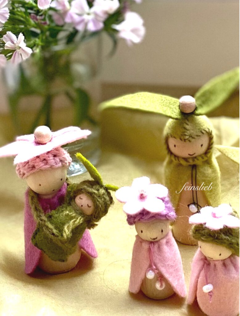Blumenelfen basteln Anleitung: Mutter, Vater und zwei Kinder mit Blumen auf dem Kopf aus Holzfiguren mit Filzumhängen, die Mutter hat ein Baby umgehängt. Auf grünem Tuch vor Blütenstrauß.