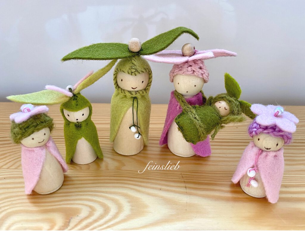 5 Holzfiguren als Blumenelfen mit Umhängen und Mützen in grün und rosa, auf Holztisch. Die Mutter hat ein Baby im Schlafsack umgehängt. Manche der Figuren haben Blumen auf den gehäkelten Mützen und sind mit 1-2 Perlen verziert.