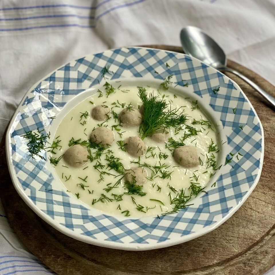 Mairübchen-Rezept: Suppenteller mit weißer Suppe und kleinen Klößchen mit viel frischem Dill darüber, auf rundem Holzbrett.
