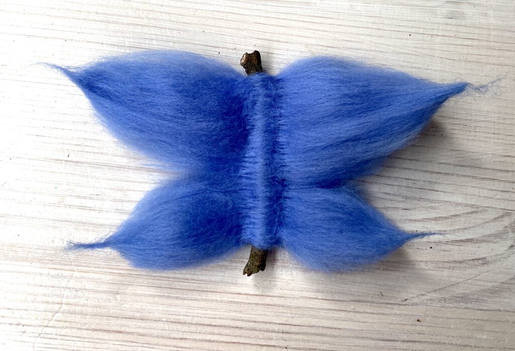 gefilzter blauer Schmetterling auf Holztisch. Der Körper ist ein Stöckchen.