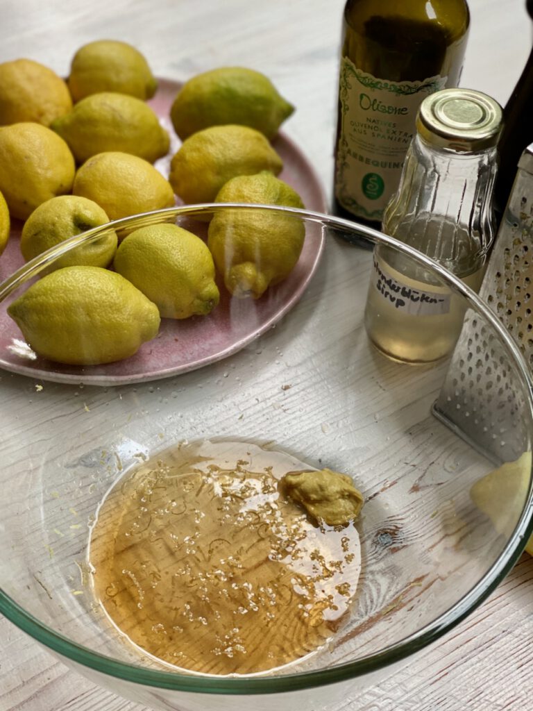Zitronen-Vinaigrette Zutaten Schüssel mit angerührter Vinaigrette, dahinter Teller mit Zitronen, Olivenöl, Holunderblüten-Sirup und Reibe mit Zitrone 