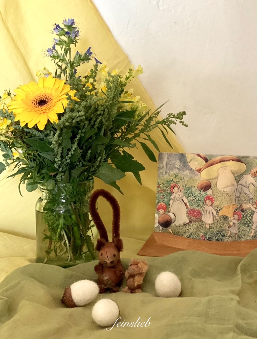 Jahreszeitentisch September mit gelbem Tuch, Vase mit Wildblumen in gelb und blau, Postkarte und kleinen Eichhörnchen aus Haselnüssen