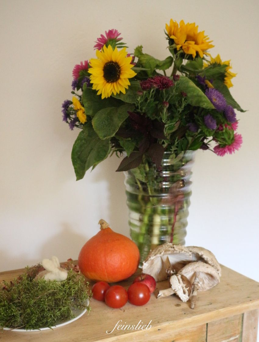 Spätsommer-Strauß mit Sonnenblumen, Tomaten, und Kürbisse und Pilze auf Holztischchen vor weißer Wand (Jahreszeitentisch September / Erntedank)