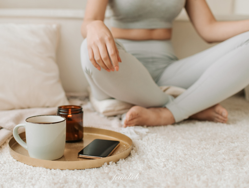 Meditation: Unterkörper einer Frau in Meditationshaltung auf Flauschteppich, vor ihr Tablett mit Teetasse, Kerze und Handy
