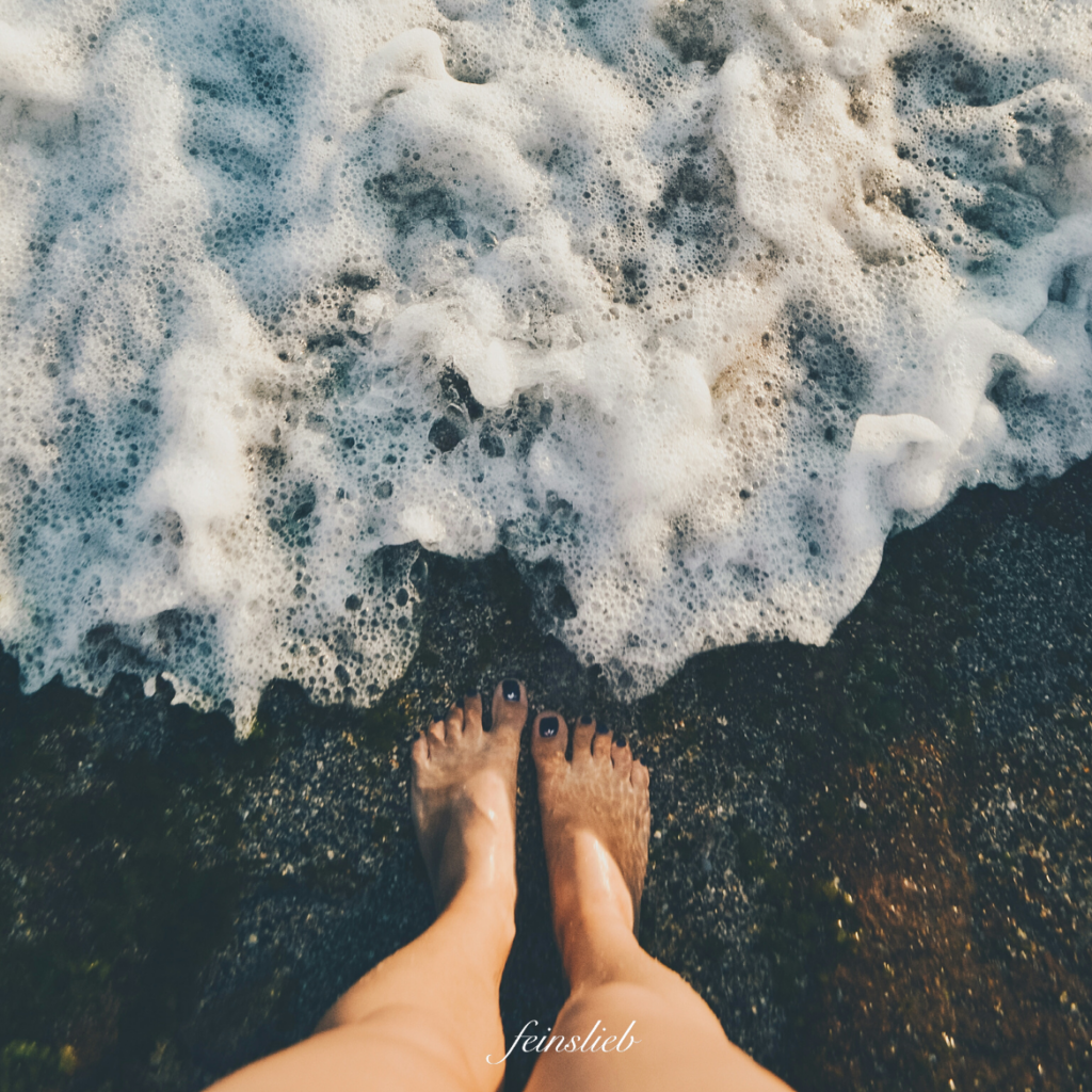 Füße am Meeressaum auf schwarzem Sand, Schaumwelle nähert sich
