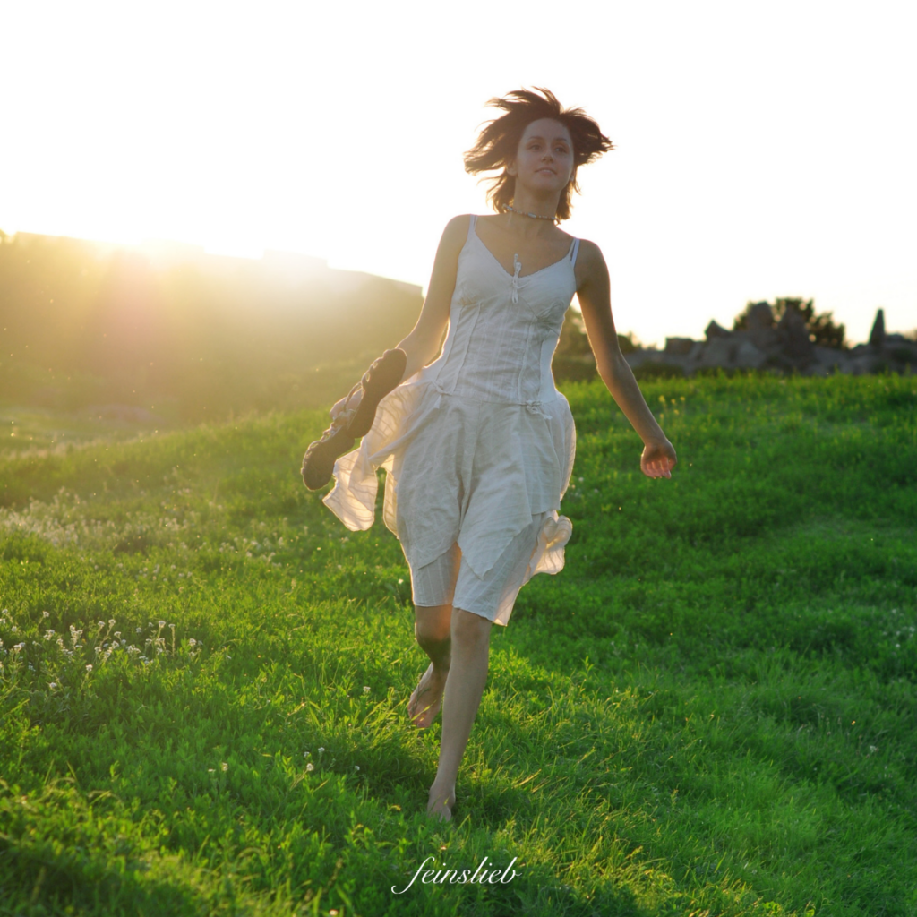 Frau im weißen Sommerkleid rennt barfuß eine Wiese entlang, die Sonne im Hintergrund