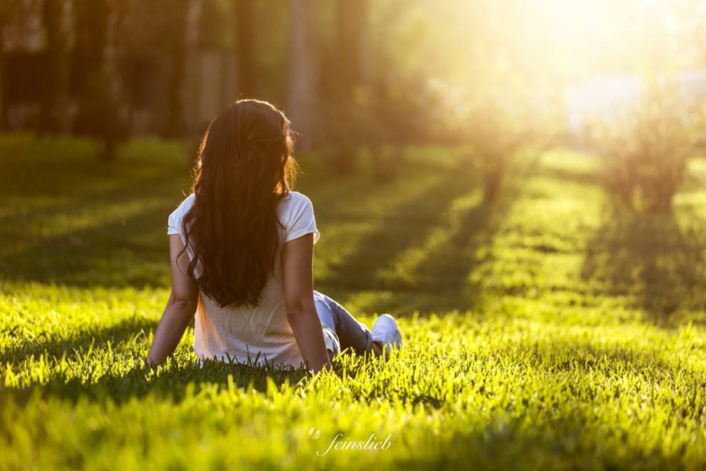 Frau mit weißem Shirt sitzt von hinten gesehen auf einer grünen Wies und schaut in die Sonne, die durch Bäume scheint