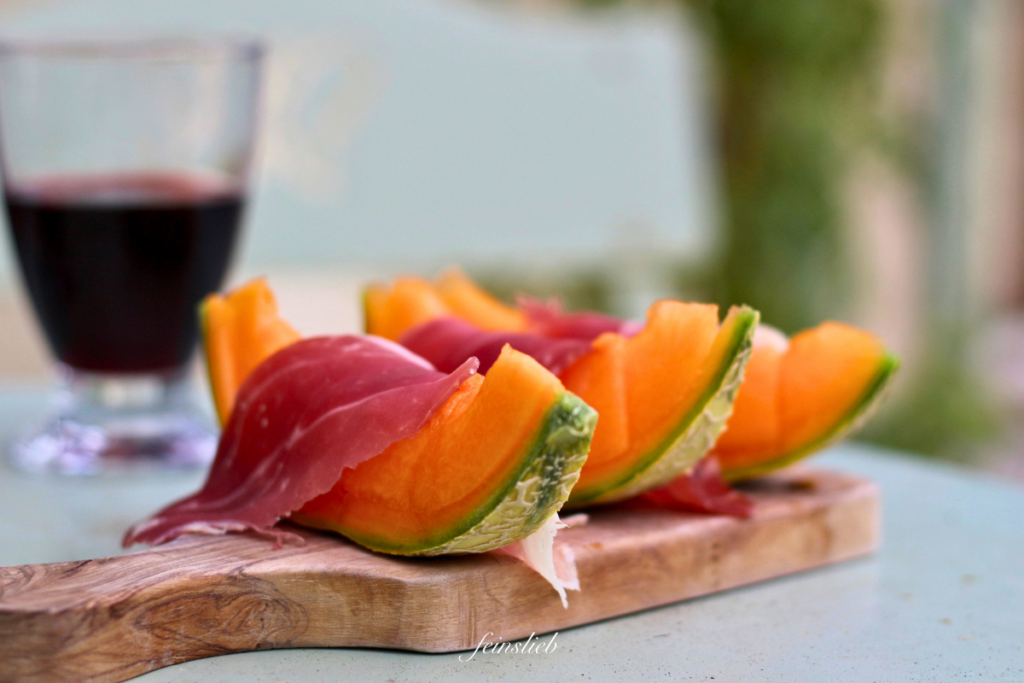 Orangene Honigmelonen-Scheiben mit Parmaschinken auf Holzbrett, Weinglas mit Rotwein dahinter